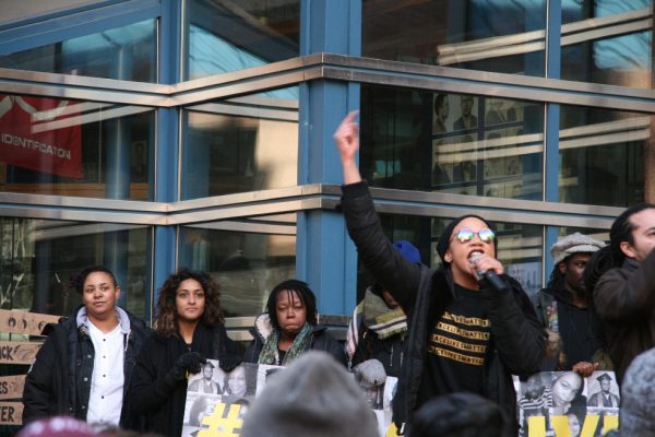 Photo of a woman with her arm in the air holding a microphone speaking to a Black Lives Matter Toronto rally on March 26